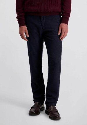 Mann trägt dunkelblaue Hose, einen burgunderfarbenen Pullover und dunkelbraune Ledermokassins und steht vor einem einfarbigen Hintergrund.