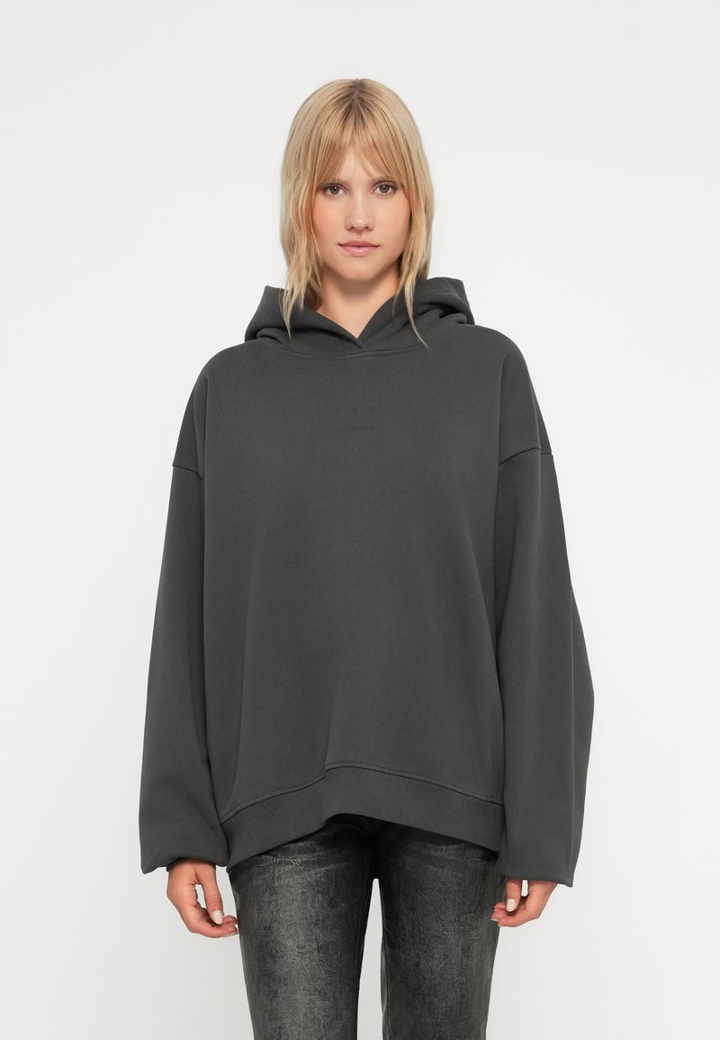 Haut à capuche ample gris foncé avec une poche kangourou, un ourlet côtelé et un capuchon ajustable, porté sur un pantalon noir à texture.