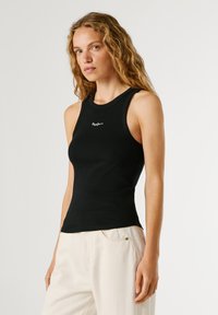 Femeie cu păr blond, lung și creț, purtând un top negru fără mâneci și pantaloni albi, stând în picioare pe un fundal alb simplu.