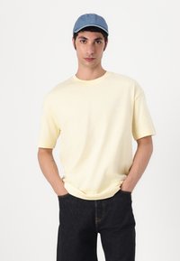 Yellow short-sleeve cotton t-shirt with a round neckline, displayed with dark jeans and a light blue denim cap. Minimal text detail on the shirt.