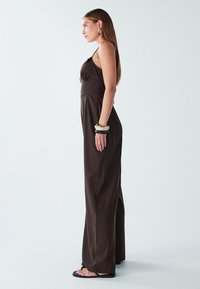 Žena stojící v profilu ve tmavě hnědém bezrukávovém kombinézónu, černých sandálech a masivních náramcích na pravém zápěstí.