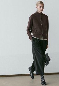 Bruine wollen cardigan met een hoge kraag en knoopsluiting aan de voorkant, gecombineerd met losse zwarte broek en zwarte enkellaarsjes. Detail van de gestructureerde stof zichtbaar.