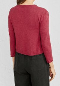 Červený, dlouhý rukáv, krčený svetr s kulatým výstřihem. Hladká textura, vroubkované manžety a lem. V kombinaci s volnými černými kalhotami se zadními kapsami.