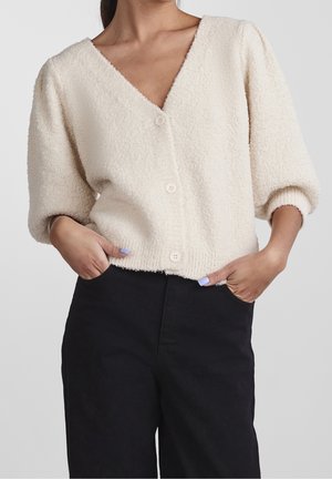 Frau trägt eine cremefarbene, strukturierte Strickjacke mit Knöpfen und Puffärmeln sowie schwarze, hoch taillierte Hosen, Hände in den Taschen.