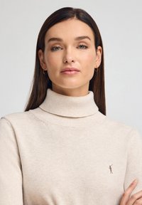 Beige coltrui gemaakt van zachte gebreide stof. Voorzien van een geribbelde kraag en een klein logo-accent op de borst. Gladde textuur.