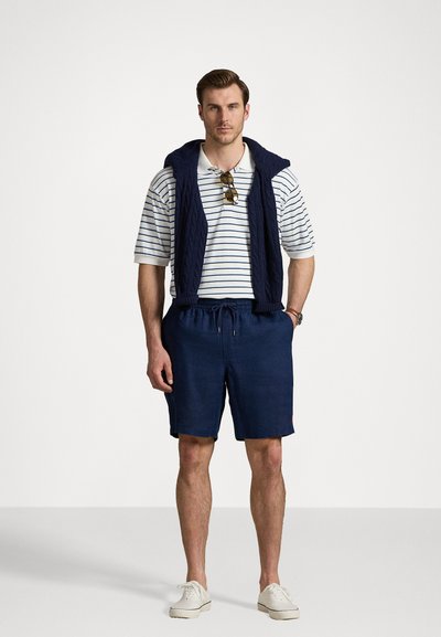 Calções azul-marinho com cordão, combinados com uma polo riscada em branco e azul-marinho. Um suéter azul-marinho canelado repousa sobre os ombros. Ténis brancos.