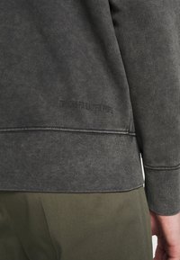 Ciemnoszara bluza z teksturowanym wykończeniem, z prążkowanym dołem i bocznymi detalami. Nazwa marki jest nadrukowana blisko dołu.