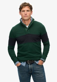 Pullover verde con una striscia orizzontale nera, colletto con mezzo zip e tessuto testurizzato. Indossato sopra una camicia di jeans, abbinato a un paio di jeans blu.