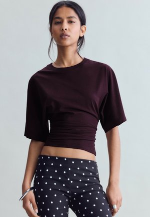 Donna in piedi con indosso una maglietta a maniche corte viola scuro e pantaloni neri con pois bianchi su uno sfondo chiaro uniforme.