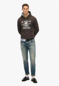 Sudadera gris oscuro con gráfico de calavera blanca y texto, jeans azules, zapatos negros y calcetines blancos. Estilo casual con un ajuste relajado.