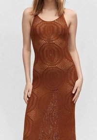 Femme portant une robe en crochet sans manches de couleur rouille avec des motifs circulaires, se tenant devant un fond clair.