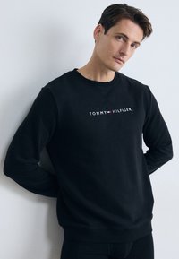 Sudadera de algodón negra con escote redondeado y puños acanalados. Texto del logo centrado en blanco "TOMMY HILFIGER," con acentos en rojo y blanco.