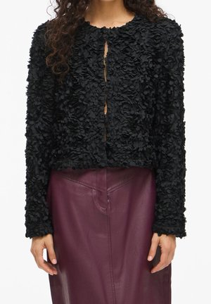 Femme portant une veste noire texturée à manches longues avec des appliques florales, associée à une jupe en cuir violet foncé taille haute.