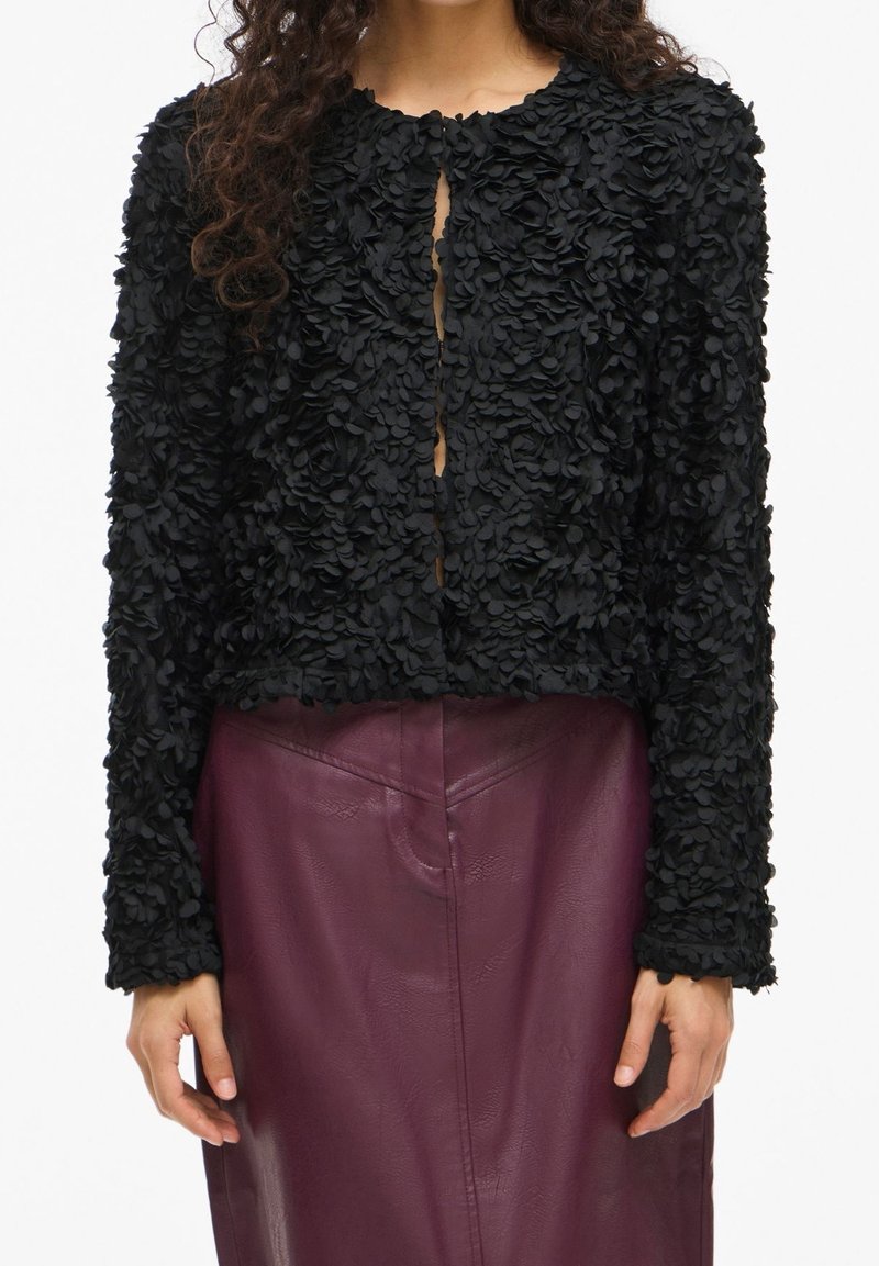 Femme portant une veste noire texturée à manches longues avec des appliques florales, associée à une jupe en cuir violet foncé taille haute.