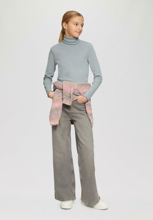 Lichtblauwe coltrui, losse grijze wijde jeans en een roze gebreid vest om de taille gebonden. Witte sneakers maken de outfit af.