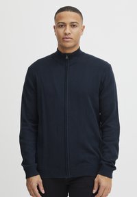 Marine blå zip-genser laget av et mykt materiale med ribbestrikket krage og mansjetter, med en subtil tekstur og en tettsittende silhuett.
