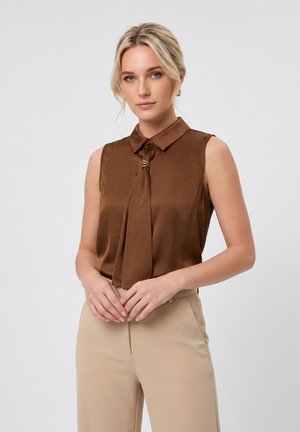 Blondynka w brązowej satynowej bluzce bez rękawów z kołnierzykiem i beżowych spodniach z wysokim stanem, stojąca z rękami splecionymi przed sobą.