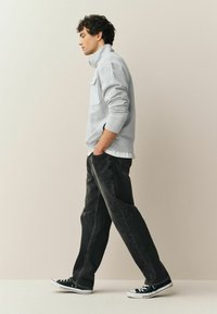 Grå sweatshirt med høj krave, sorte denimjeans og sorte canvas-sneakers. Modellen står i profil mod en neutral baggrund.