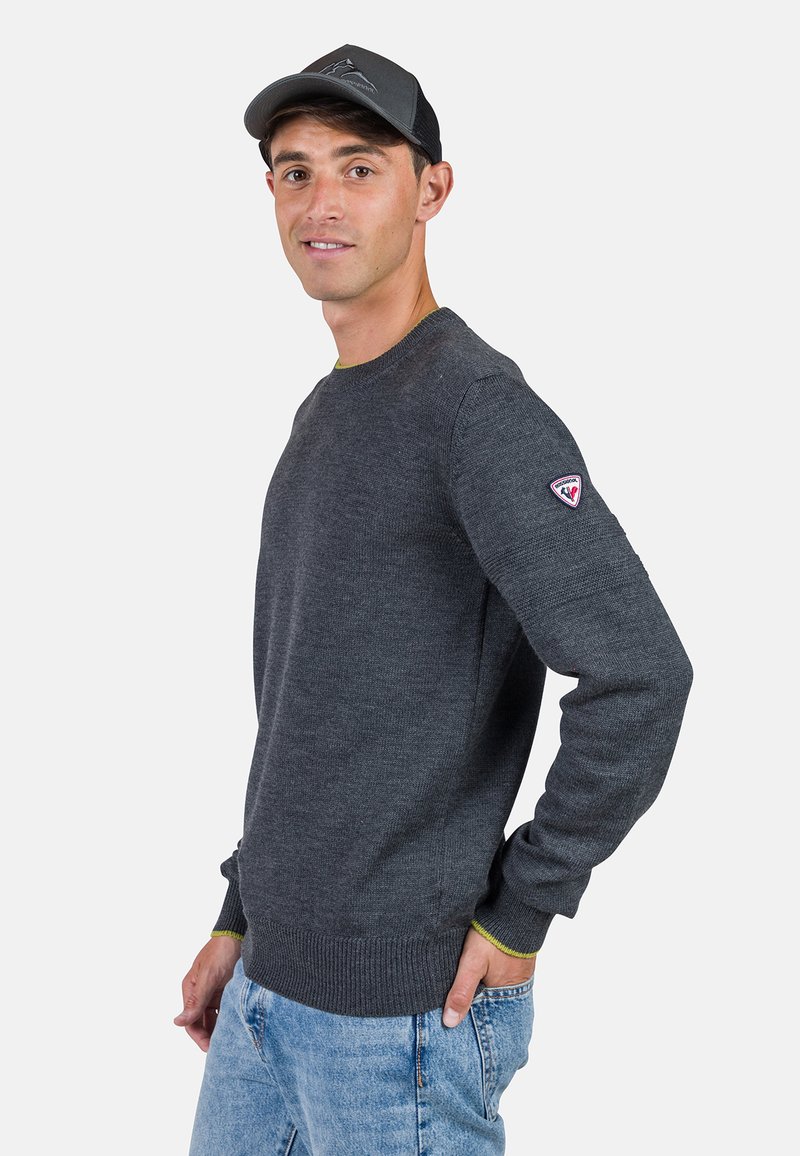 Dunkelgrauer gestrickter Pullover mit Rundhalsausschnitt, enganliegenden Bündchen und Saum; mit einem dezenten grünen Akzent entlang des Kragens; getragen mit blauen Jeans.