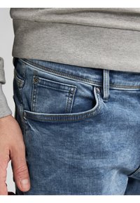 Jeans in denim blu sbiadito con cinque tasche, cuciture visibili e una finitura testurizzata. Maniche di una felpa grigia parzialmente visibili in alto.