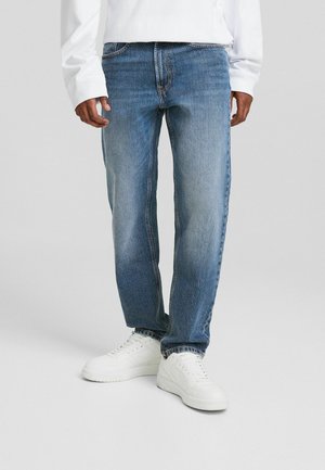 Personne portant un jean bleu et des baskets blanches debout devant un fond clair uni.