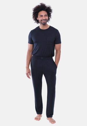 Uomo con capelli ricci in piedi a piedi nudi, indossa una camicia nera a maniche corte e pantaloni abbinati, sorride e guarda di lato.