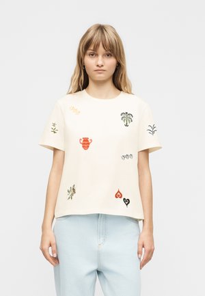 Mladá žena v krémovom tričku s krátkym rukávom s farebnými vyšívanými motívmi a svetlomodrých džínsoch s vysokým pásom, stojaca pred jednoduchým pozadím.