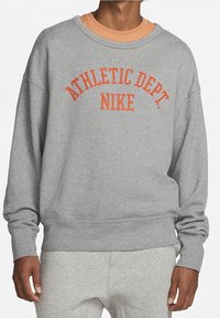 Sudadera gris de cuello redondo con texto naranja que dice "DEPARTAMENTO ATLÉTICO NIKE," con puños y dobladillo acanalados. Lleva puesta una camiseta de color naranja claro debajo.