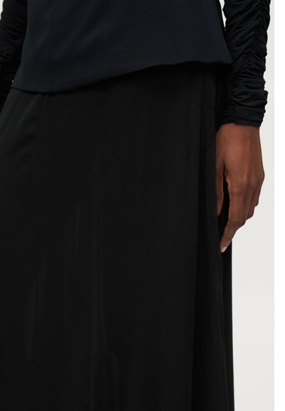 Højre hånd hvilende ved siden iført en sort ruched top med lange ærmer og sort nederdel mod en lys baggrund.