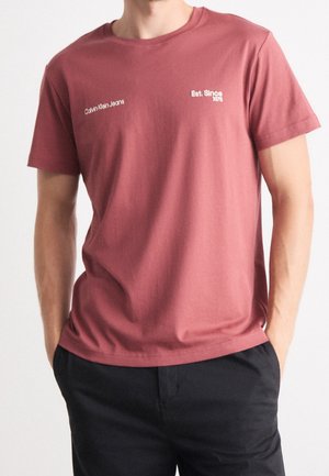 Mann trägt ein gedämpft rotes Calvin Klein Jeans T-Shirt mit weißem Text "Calvin Klein Jeans" und "Est. Since 1978," kombiniert mit schwarzen Hosen.