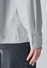Camicia a maniche lunghe grigia chiara realizzata in tessuto texture, con polsini abbottonati e uno spacco laterale, abbinata a jeans scuri.