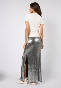 Srebrna spódnica maxi z cekinami i bocznymi rozcięciami, zestawiona z kremowym topem z krótkim rękawem. Widoczna jest gładka tekstura i mieniące się powierzchniowe detale.