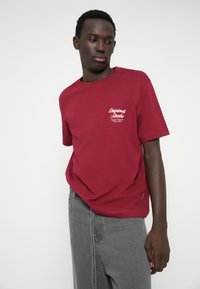 Burgundijas kokvilnas t-kreklis ar īsām piedurknēm un apaļu kakla izgriezumu, ar baltu teksta dizainu kreisajā krūšu daļā; apvienots ar pelēkiem biksēm.