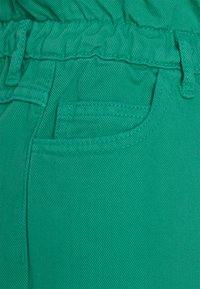 Tecido verde texturizado de um bolso de calças com detalhes de costura visíveis. Cintura elástica com um design franzido e passantes para cinto.