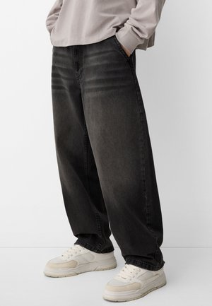 Persoon draagt vervaagde zwarte wijde jeans, beige sweatshirt en witte sneakers met beige accenten, staat met handen in de zakken.