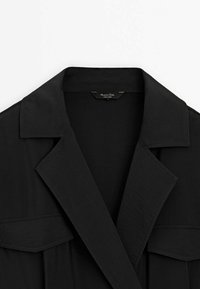 Schwarzes Hemd mit breiten Kragen und zwei Brusttaschen mit Klappen, flach auf einem weißen Hintergrund gelegt.