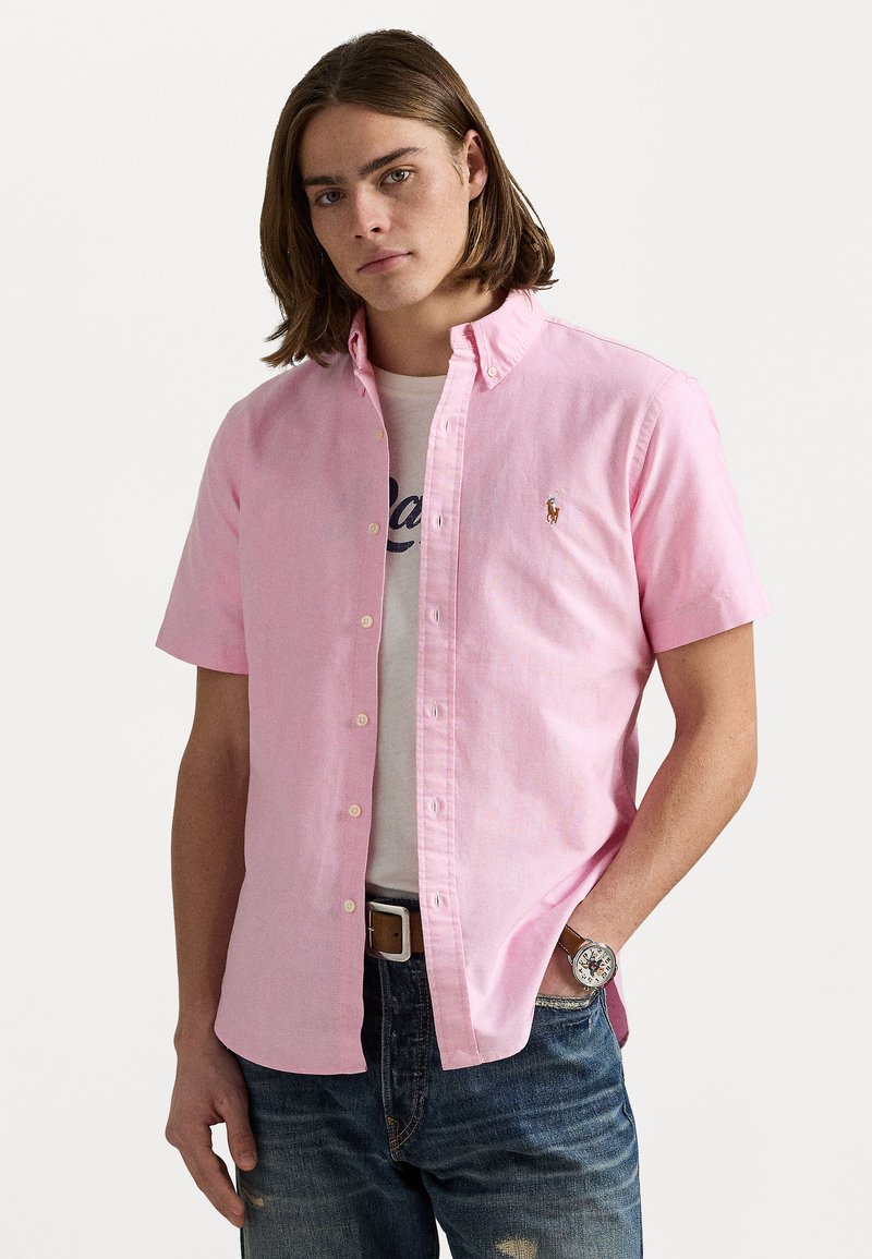Chemise rose clair à manches courtes avec col boutonné, arborant un petit logo brodé. Portée par-dessus un t-shirt blanc et associée à un jean en denim.