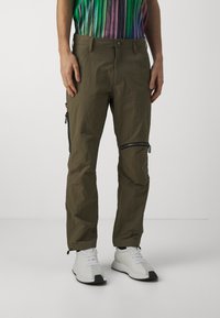 Cargo nohavice v olivovej zelenej farbe, vyrobené z ľahkého materiálu. Disponujú vreckami na zips a zúženými nohavicami, skombinované s bielymi teniskami.