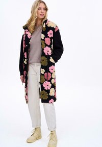 Dlhý čierny kabát so širokými ružovými, krémovými a zelenými kvetinovými vzormi, s kapucňou, prednými gombíkmi a bočnými vreckami. Nosený cez šedý top.