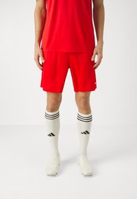 Tricou sportiv roșu și pantaloni scurți, cu șosete cu dungi albe și negre. Pantofii de fotbal sunt de o culoare deschisă, cu o textură netedă și un design ajustat.