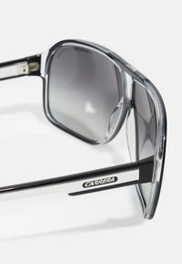 Ochelari de soare cu lentile gradiente de gri închis, rame groase transparente și negre, având un accent argintiu și logo-ul "Carrera" pe lateral.