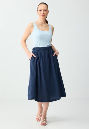 Femme portant un haut sans manches bleu clair et une jupe midi bleu marine, les mains dans les poches, chaussée de sandales blanches à talons, sur fond uni.