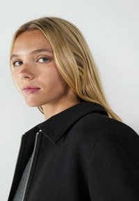 Schwarze Jacke mit strukturiertem Kragen und silbernem Reißverschluss. Glatte Textur, minimalistisches Design, taillierte Form. Teilweise sichtbares blondes Haar.