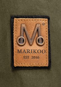 Brun läderlapp med präglad logotyp "M" och texten "MARIKOO EST 2016" på en olivgrön tygbakgrund med svarta sömnadsdetaljer.