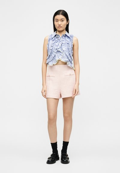 Γυναίκα που φοράει μπλε κοντό τοπ με βολάν χωρίς μανίκια, ροζ υφασμάτινη ψηλόμεση βερμούδα, μαύρες κάλτσες και μαύρες μοκασίνια, στέκεται μπροστά σε λευκό φόντο.