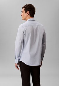 Hele sinise tooniga pika varrukaga särk, millel on kraega kaelus, nööbitavad mansetid ja ümar serv, kantud tume värvi pükstega. Sile kangapind.