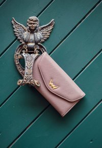 Pink textured wallet with gold accents, featuring a flap closure, displayed against a green wooden background with a decorative metal hook.