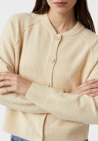 Kremowy kardigan o miękkiej fakturze, z prążkowanym kołnierzem i mankietami, zapinany na pięć guzików, o luźnym kroju.