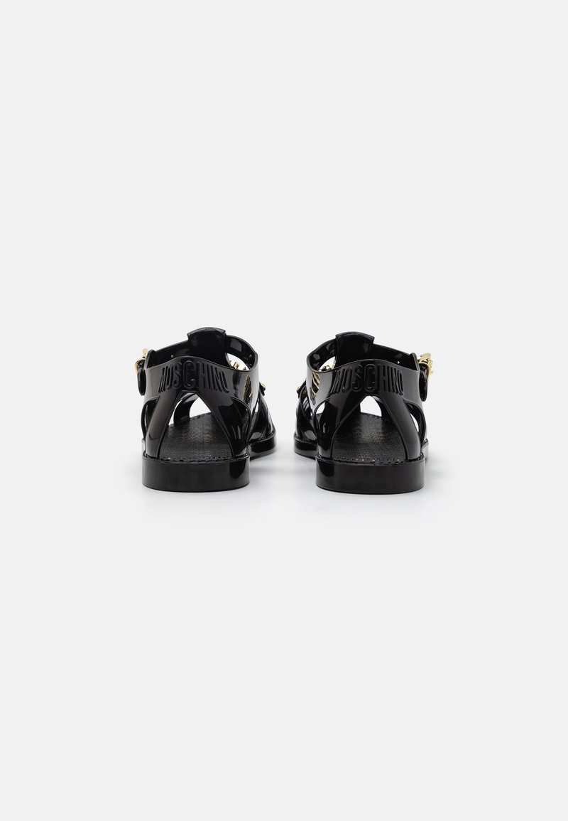 Sandales en cuir verni noir avec un design découpé, une sangle de cheville réglable et un logo en relief. Semelle plate avec un minimum de détails.