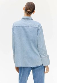 Giacca di jeans blu chiaro con vestibilità rilassata, cuciture visibili, maniche lunghe e polsini con bottoni. Il tessuto ha una texture liscia.